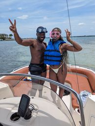 Two people on a motorboat wearing snorkel masks and a blue life jacket, flashing peace signs with clear coastal bay waters and a sunny sky in the background