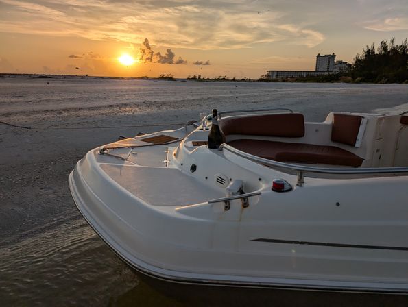 White motorboat pulled up on a sandy beach at sunset with a champagne bottle on the bow, calm shallow water and a golden sky, distant shoreline buildings and trees