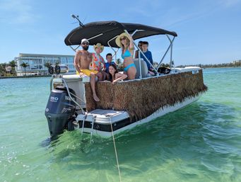 Adults and children on a tiki-trimmed pontoon boat floating in clear turquoise coastal water on a sunny day, shoreline buildings and palm trees in the background.