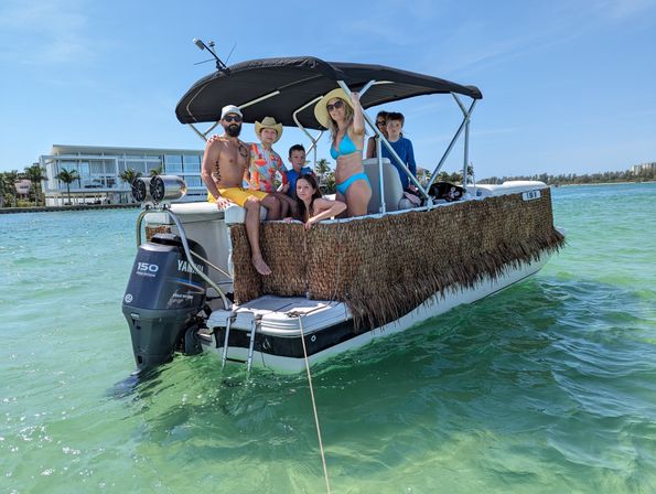 Adults and children on a tiki-trimmed pontoon boat floating in clear turquoise coastal water on a sunny day, shoreline buildings and palm trees in the background.