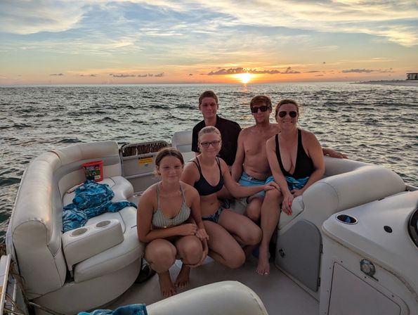 Smiling group of five in swimsuits on a white pontoon boat at sunset, colorful sky and sun dipping over open water with gentle waves on the horizon.