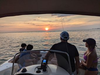 Group on a motorboat watching a golden ocean sunset from the bow, with a fishing rod and fishfinder visible on the console.