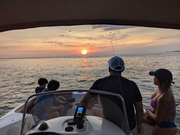Group on a motorboat watching a golden ocean sunset from the bow, with a fishing rod and fishfinder visible on the console.