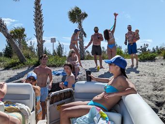 Group of friends enjoying a sunny beach day on a boat and sandy shoreline with palm trees—people in swimsuits chatting, drinking, and playing near the water.