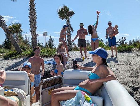 Group of friends enjoying a sunny beach day on a boat and sandy shoreline with palm trees—people in swimsuits chatting, drinking, and playing near the water.