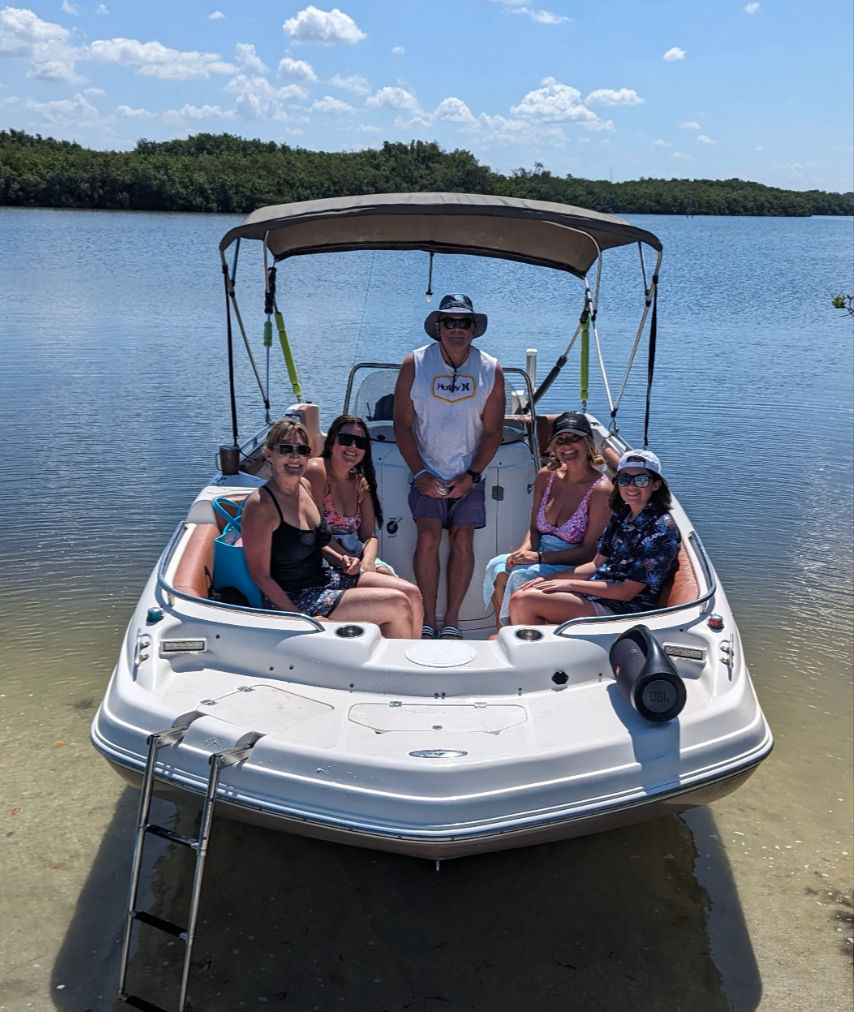 Five people relaxing on a white motorboat with a bimini top, anchored in shallow clear water near a mangrove-lined inlet on a sunny blue-sky day, swim ladder and portable speaker visible.