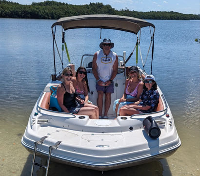 Five people relaxing on a white motorboat with a bimini top, anchored in shallow clear water near a mangrove-lined inlet on a sunny blue-sky day, swim ladder and portable speaker visible.