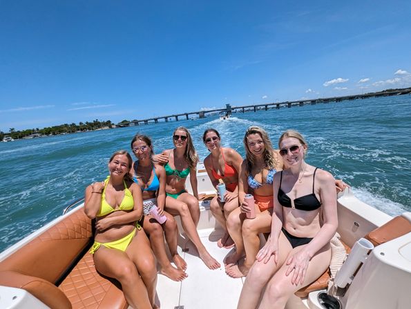 Six women in bright bikinis smiling on a motorboat with tan quilted seats, cruising on blue coastal waters past a low bridge under a clear sunny sky