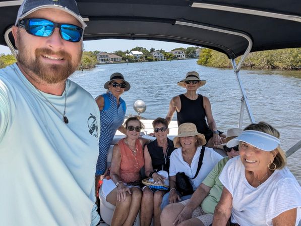Group of smiling adults on a covered motorboat enjoying a sunny coastal waterway lined with mangroves and waterfront homes, many wearing sun hats, visors and sunglasses.