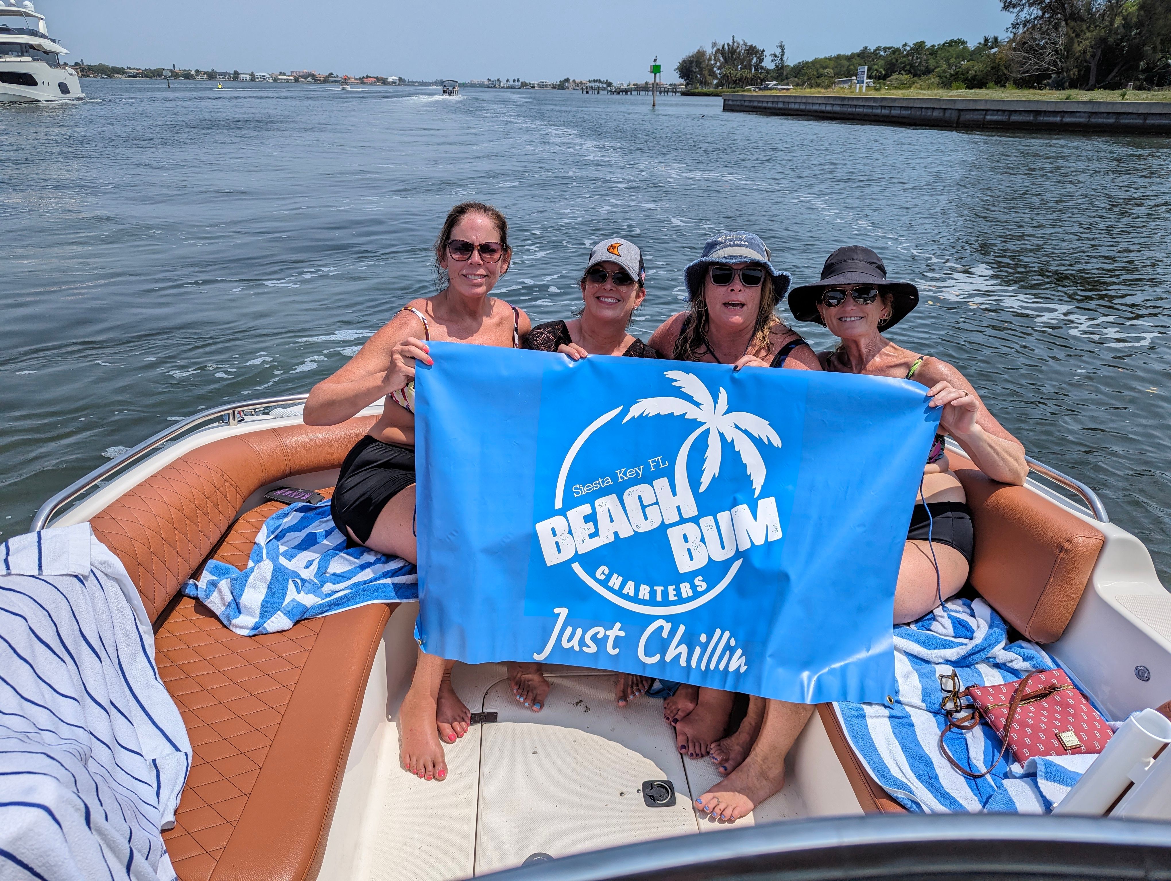 Four women smiling on a motorboat near Siesta Key, Florida, holding a bright blue banner with a palm-tree graphic and the slogan Just Chillin on calm coastal waters.
