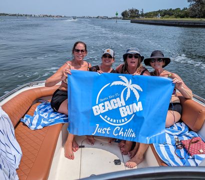 Four women smiling on a motorboat near Siesta Key, Florida, holding a bright blue banner with a palm-tree graphic and the slogan Just Chillin on calm coastal waters.