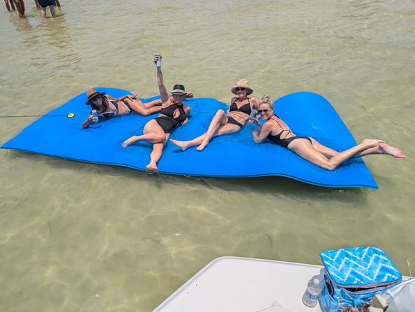 Four friends lounging on a bright blue floating mat in shallow clear water near a boat, wearing swimsuits and sun hats, holding drinks and soaking up the sun.