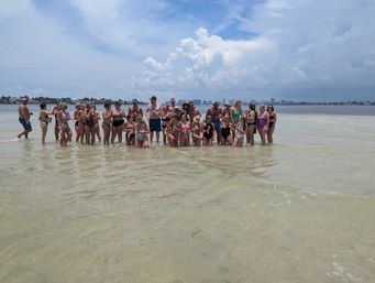 Large group of people in swimsuits gathered on a shallow sandbar in calm coastal water, posing on a sunny summer day with puffy clouds and a distant city skyline in the background.
