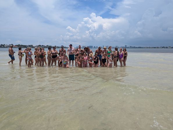 Large group of people in swimsuits gathered on a shallow sandbar in calm coastal water, posing on a sunny summer day with puffy clouds and a distant city skyline in the background.
