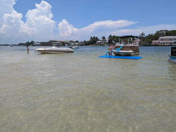 Shallow clear water at a sunny coastal sandbar with anchored leisure boats and a person climbing from a blue floating mat onto a pontoon, palm trees and waterfront homes under puffy summer clouds.