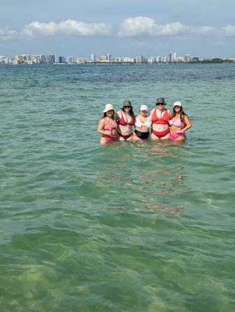 Group of five women in colorful bikinis and sun hats standing waist-deep in clear green coastal bay water with a distant urban skyline and sunny sky