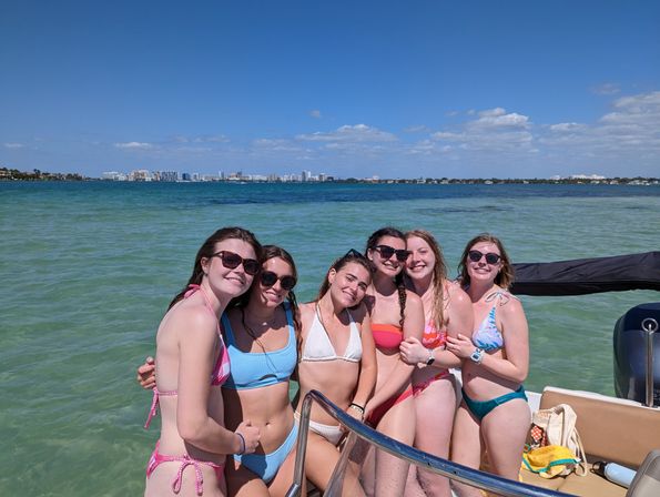 Group of six friends in colorful bikinis posing on a boat in clear turquoise bay on a sunny day, with a distant coastal city skyline and blue sky.