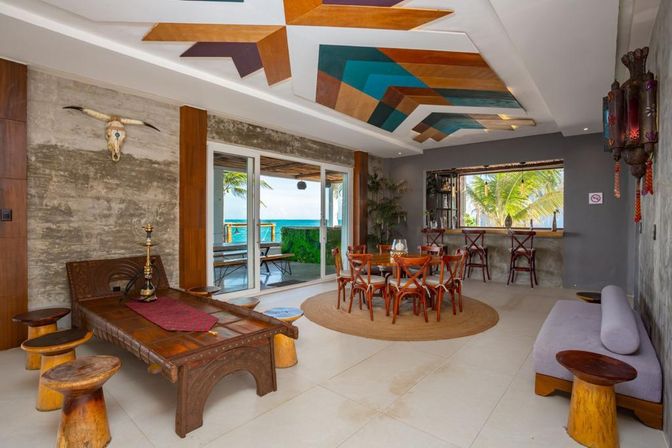 Beachfront tropical lounge and dining room with rustic wooden furniture, round dining table, bar seating, colorful geometric ceiling panels and sliding glass doors framing ocean and palm trees.