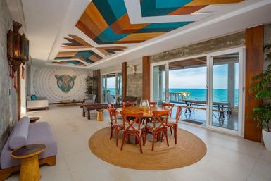 Oceanfront boho-modern dining and lounge with round wooden table on a woven rug, vibrant teal-and-amber geometric ceiling mural, decorative animal wall mosaic, and sliding glass doors opening to a seaside deck and turquoise ocean view.