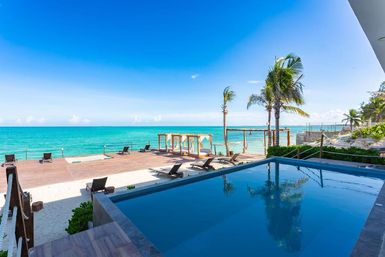 Tropical beachfront scene with a private infinity pool reflecting palm trees, wooden deck with cabanas and lounge chairs overlooking turquoise ocean and clear blue sky.