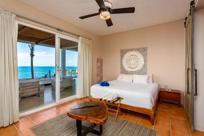 Oceanfront bedroom with king bed, sliding glass doors opening to a beachfront patio and palm-tree sea view, terracotta tile floor, rustic live-edge wood table and neutral coastal decor.