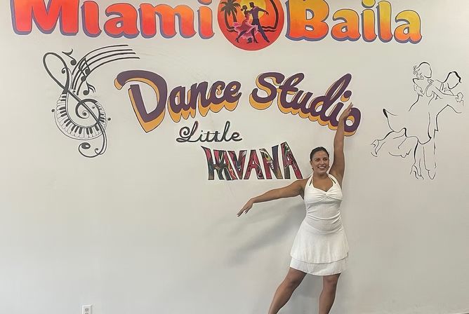 Smiling dancer in a white dress striking a graceful pose against a colorful dance studio mural with musical notes and silhouetted dancers.