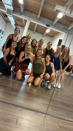 Smiling group selfie of a dozen women in a dance/fitness studio — kneeling and standing in front of a large mirror on a hardwood floor with exposed-beam ceiling and industrial lighting, post-class vibe.