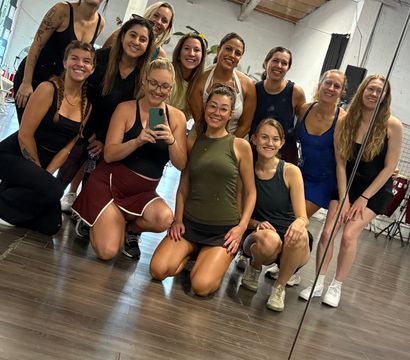 Smiling group selfie of a dozen women in a dance/fitness studio — kneeling and standing in front of a large mirror on a hardwood floor with exposed-beam ceiling and industrial lighting, post-class vibe.