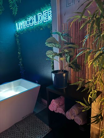 Cozy neon-lit cold-plunge spa corner with glowing blue sign, white plunge tub, potted tropical plants, bamboo screen and neatly rolled towels