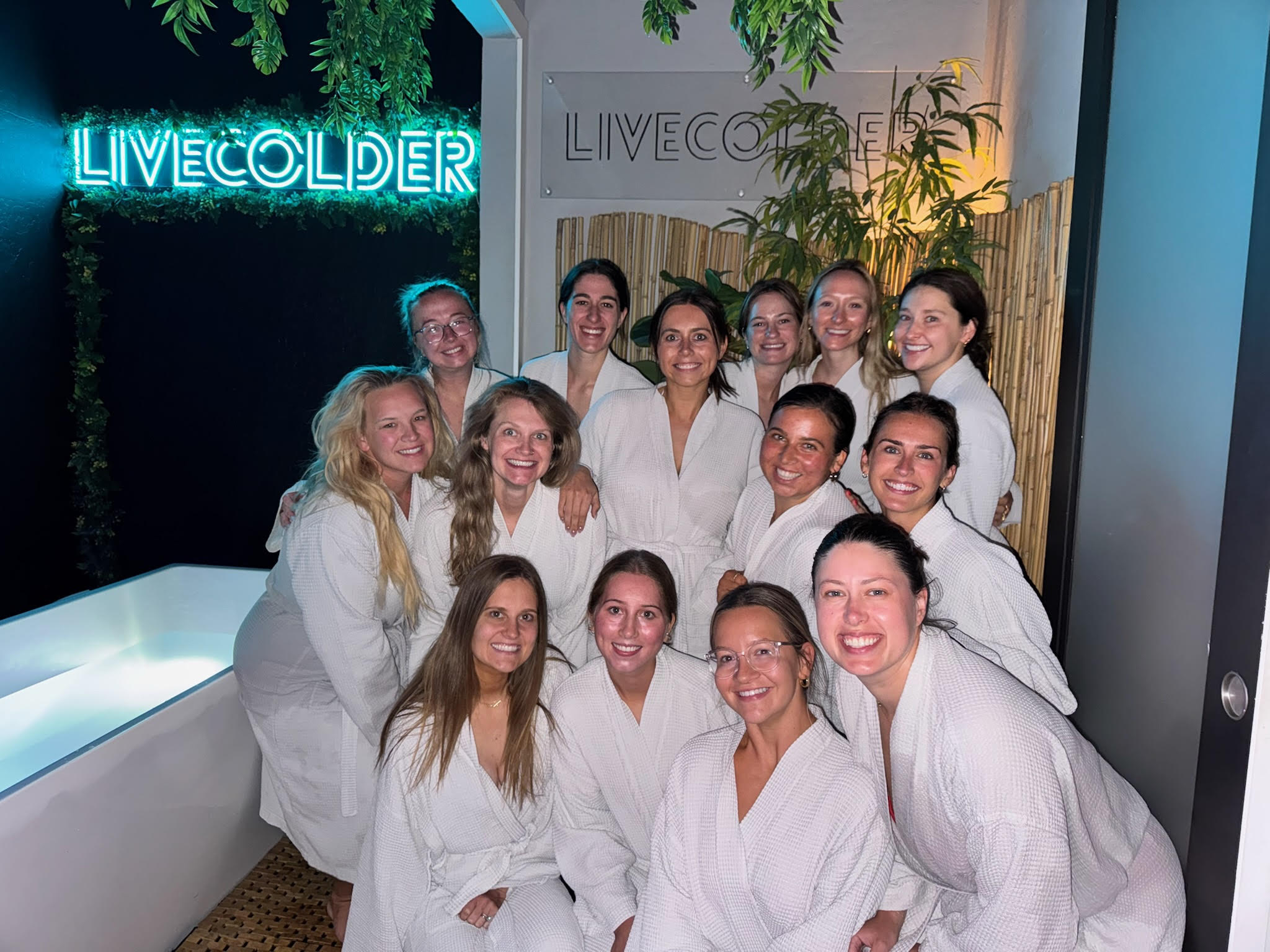 Smiling group of women in white spa robes posing in a dimly lit wellness lounge with a glowing blue neon sign, bamboo and hanging plants, and a cold-plunge tub