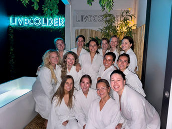 Smiling group of women in white spa robes posing in a dimly lit wellness lounge with a glowing blue neon sign, bamboo and hanging plants, and a cold-plunge tub
