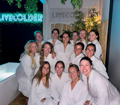 Smiling group of women in white spa robes posing in a dimly lit wellness lounge with a glowing blue neon sign, bamboo and hanging plants, and a cold-plunge tub