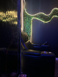 Black leather recliner in a dim indoor lounge with LED-lit living green wall, curving neon strip lights, reflective water panel and a small pebble planter.