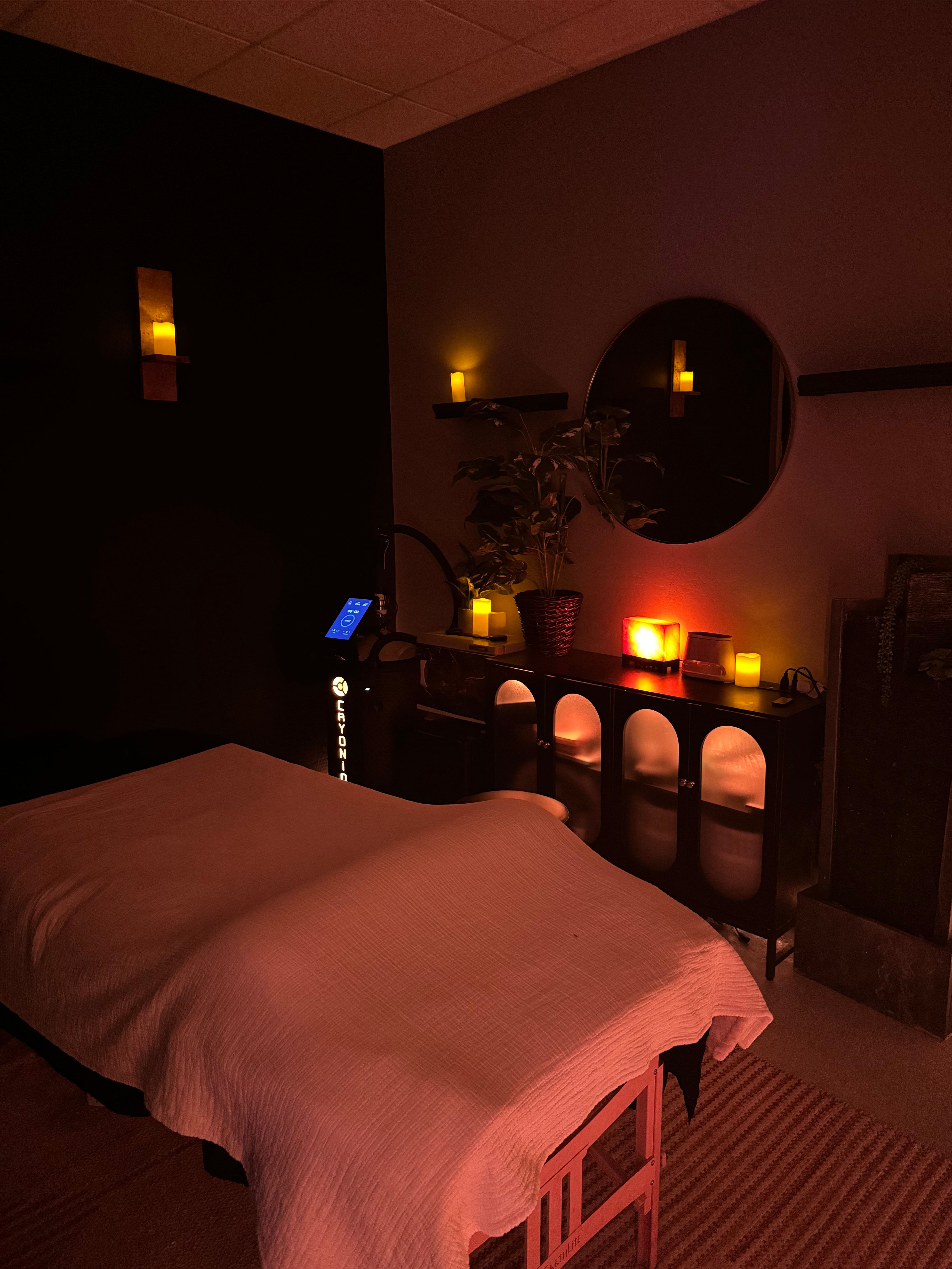 Cozy candlelit spa massage room with a covered treatment table, warm salt-lamp glow, round wall mirror, potted plant and soft ambient lighting.