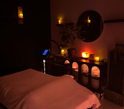 Cozy candlelit spa massage room with a covered treatment table, warm salt-lamp glow, round wall mirror, potted plant and soft ambient lighting.