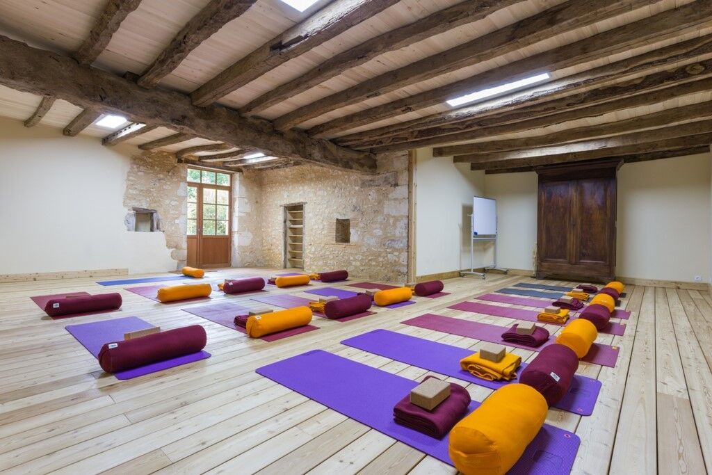 Rustic yoga studio interior with exposed wooden beams and stone walls, natural light filling a bright hardwood-floor room with rows of purple mats, burgundy bolsters and orange blankets neatly arranged for a class