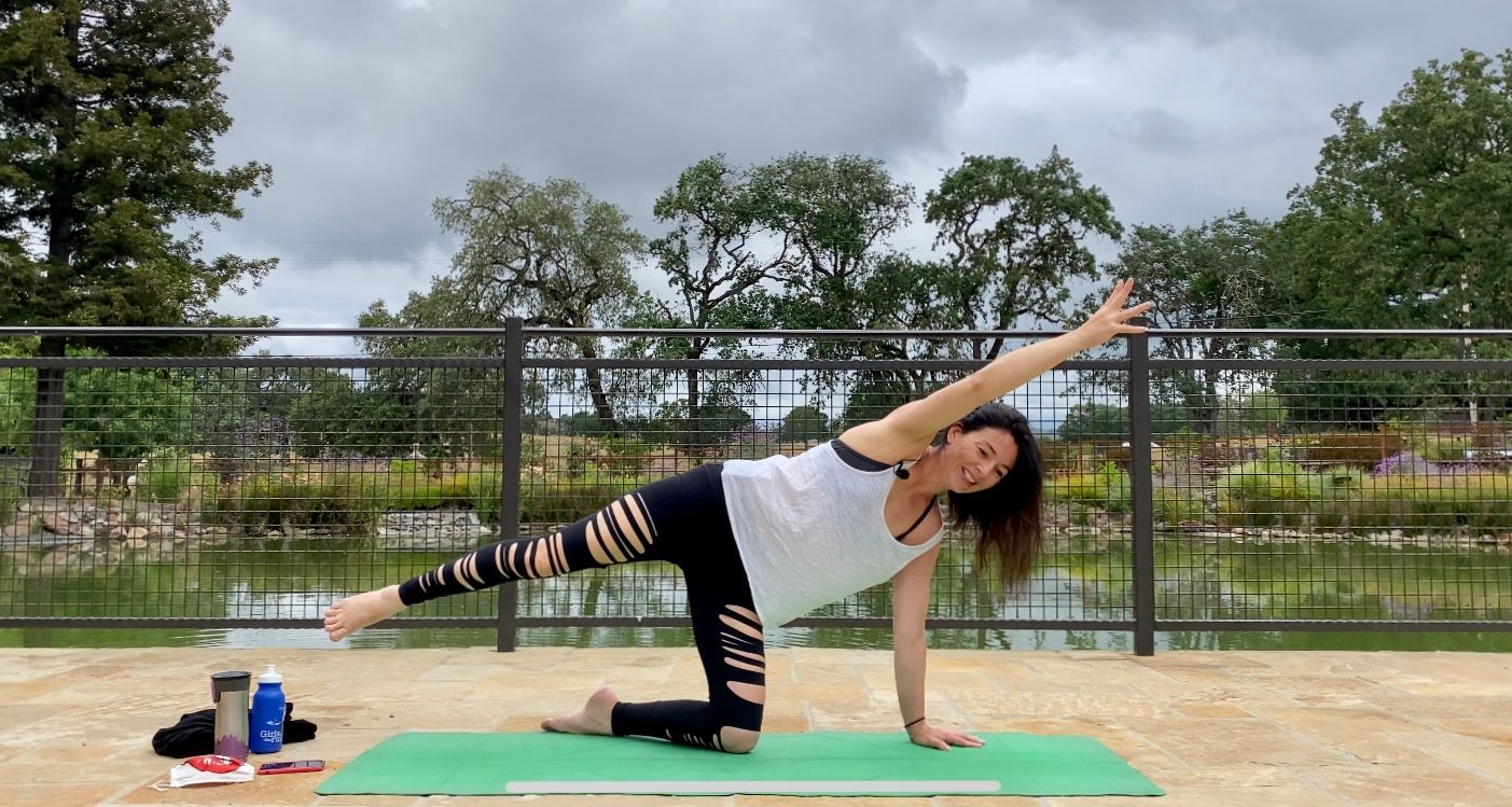 Person smiling while doing a side-kick yoga pose on a green mat on a stone lakeside patio, with metal railing, pond and oak trees under a cloudy sky; water bottle and towel beside the mat.