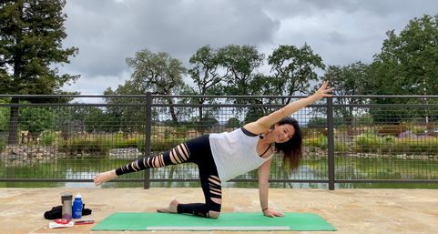 Person smiling while doing a side-kick yoga pose on a green mat on a stone lakeside patio, with metal railing, pond and oak trees under a cloudy sky; water bottle and towel beside the mat.