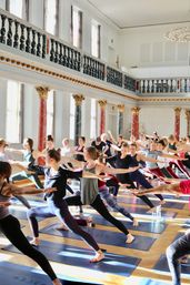 Sunlit yoga class in an ornate neoclassical hall with marble columns and balcony, dozens practicing Warrior II on mats.