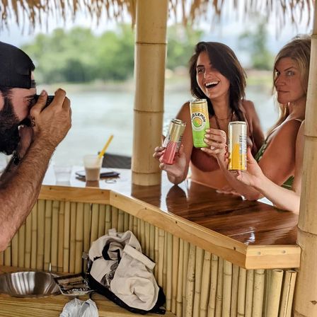 Two women in swimsuits at a bamboo tiki bar hold colorful canned drinks toward a photographer by a calm lakeside.