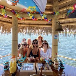 Smiling group of friends under a thatched floating tiki hut on a sunny lake, holding colorful cocktails at a wooden table decorated with leis, party boxes, a bell and a bottle.