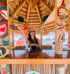 Smiling person in sunglasses and a straw visor steering a wooden wheel at a waterfront bamboo tiki hut bar, with colorful carved tiki poles, an American flag and ocean shoreline in the background