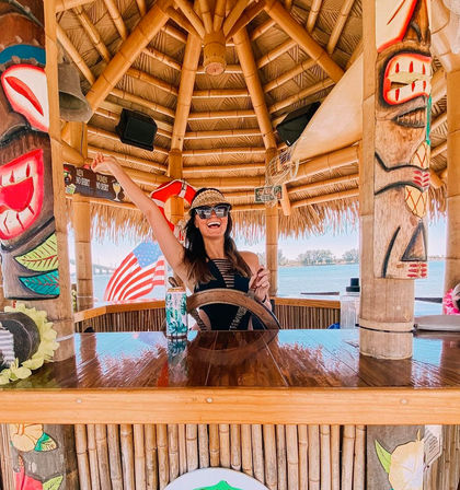 Smiling person in sunglasses and a straw visor steering a wooden wheel at a waterfront bamboo tiki hut bar, with colorful carved tiki poles, an American flag and ocean shoreline in the background