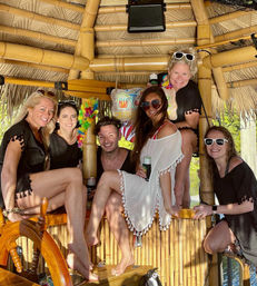 Six friends smiling in a bamboo tiki hut on a sunny tropical waterfront boat, wearing sunglasses and cover-ups and holding canned drinks.