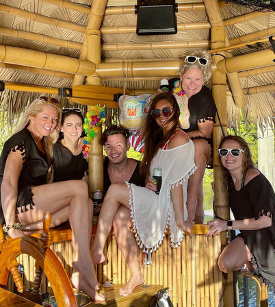Six friends smiling in a bamboo tiki hut on a sunny tropical waterfront boat, wearing sunglasses and cover-ups and holding canned drinks.