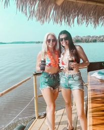 Two friends in bikinis and denim shorts wearing leis and sunglasses, holding cold drinks on a tiki-style lakeside dock under a thatched roof on a sunny summer day