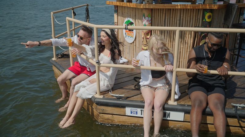 Four friends sitting on the edge of a floating tiki bar dock, feet dangling in the water, enjoying summer drinks and laughter