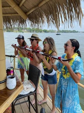 Group of four people in leis and sunglasses on a lakeside tiki deck, laughing and taking shots from a long wooden shot-ski under a thatched roof with boat and shoreline in the background.