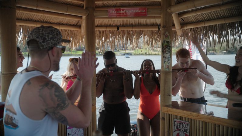 Group of friends at a floating bamboo tiki bar on a lake taking a communal shot from a long wooden shot-ski under a thatched roof, people in swimsuits cheering.