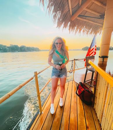 Woman in sunglasses and green lei standing on a wooden tiki-boat deck at lake sunset, holding a drink with an American flag in the background.